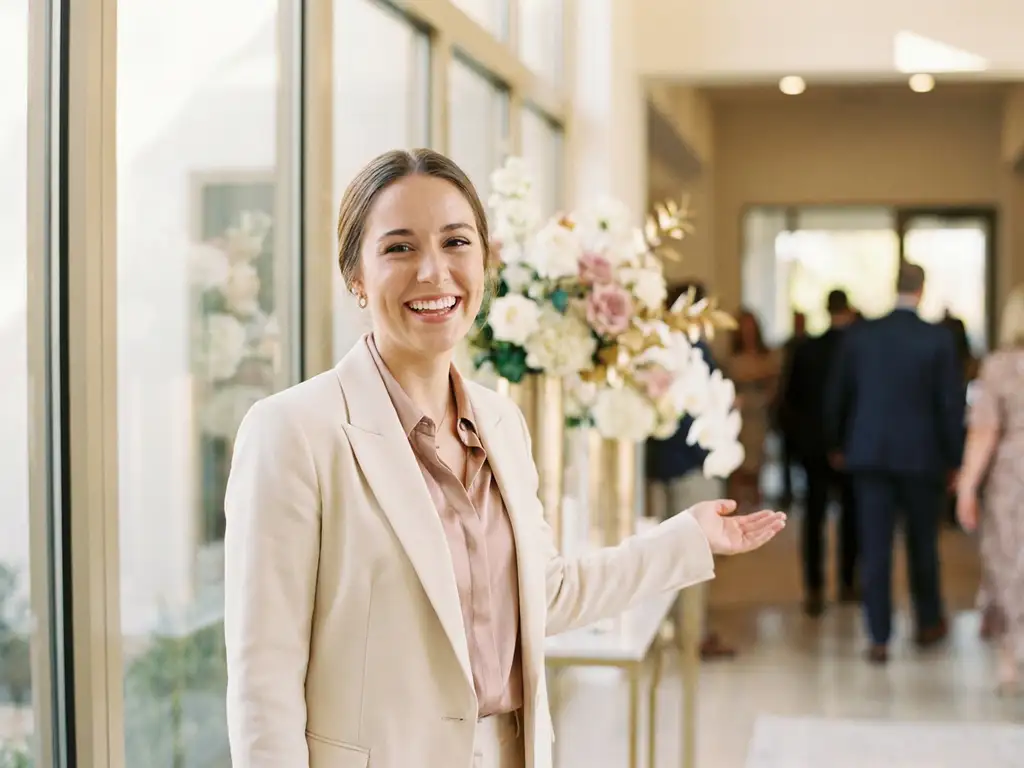 Elegante Empfangsdame begrüßt Gäste am Eingang eines modernen Veranstaltungsorts mit natürlichem Licht und floralen Arrangements.