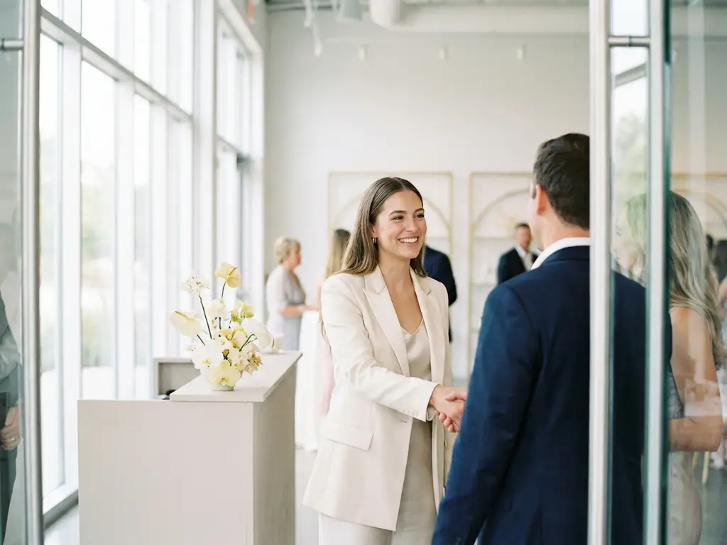 Junge Frau im hellen Blazer begrüßt Gäste am Eingang eines modernen Event-Venues mit natürlichem Licht und floraler Rezeptionsdeko.